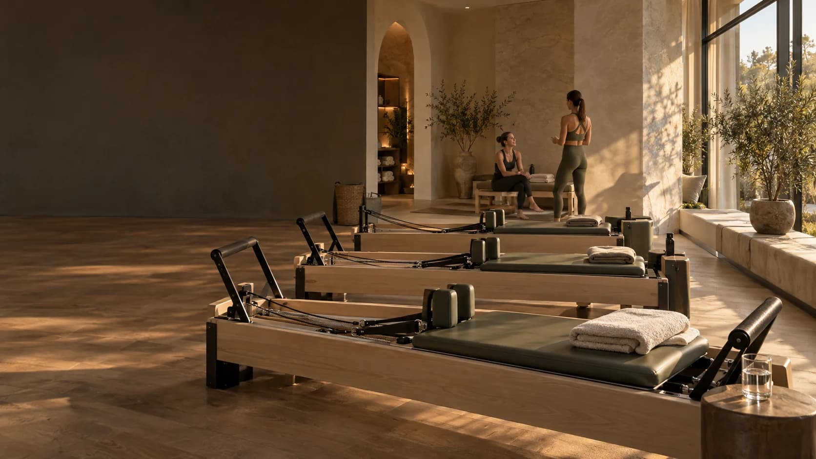 Sunlit Pilates studio with timber reformers and an instructor talking with a customer; illustrative studio image